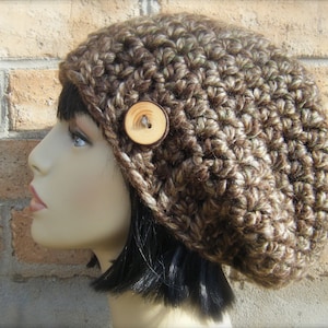 Puede incluir: Un gorro de punto marrón y beige con un botón de madera. El gorro es holgado y tiene un ajuste holgado.