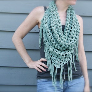 May include: A crocheted, open-weave scarf in a soft, muted teal color. The scarf features a triangular shape with fringe along the bottom edge. The open weave design creates a delicate, airy look. The scarf is worn around the neck and shoulders.