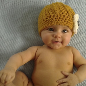 May include: A baby wearing a yellow crocheted hat with a white flower accent. The hat has a textured pattern and the baby is looking at the camera.