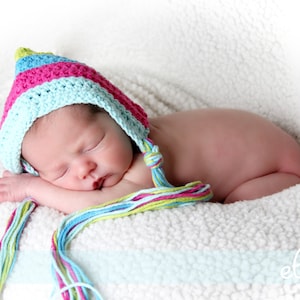 May include: A sleeping baby wearing a colourful crocheted hat with a long tassel. The baby is lying on a white fluffy blanket.