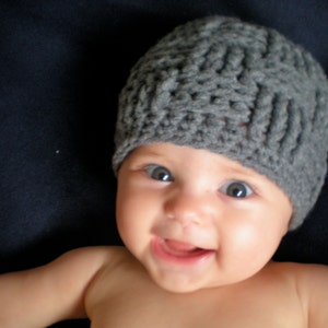 May include: A baby wearing a gray knitted hat with a simple stitch pattern. The baby has blue eyes and is smiling.
