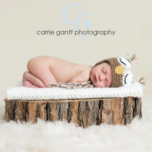 May include: A newborn baby wearing a brown and white owl-themed knitted hat is sleeping on a white blanket on a wooden log. The baby is lying on its side with its head resting on the blanket. The background is a light beige color.