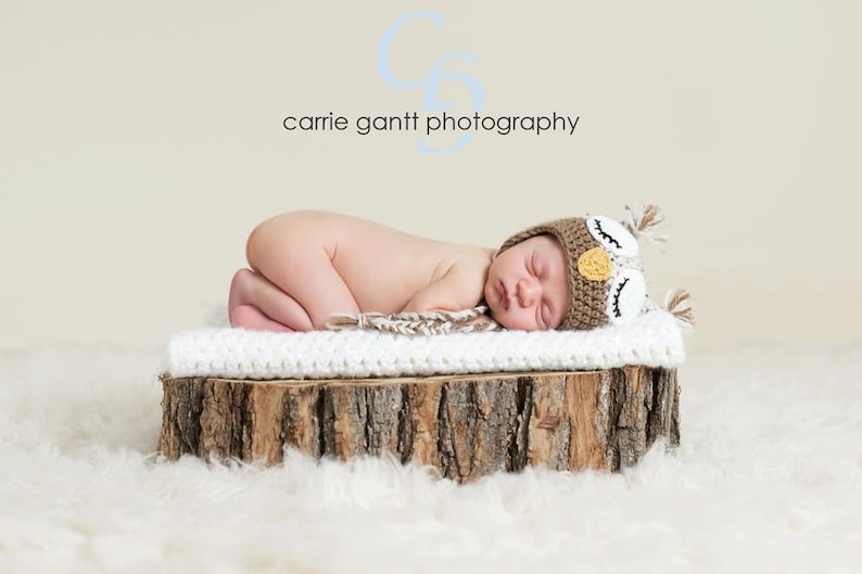 May include: A newborn baby wearing a brown and white owl-themed knitted hat is sleeping on a white blanket on a wooden log. The baby is lying on its side with its head resting on the blanket. The background is a light beige color.