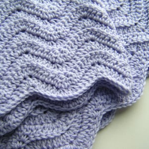 May include: A close-up of a crocheted blanket in a light purple color. The blanket has a scalloped edge and a textured pattern.