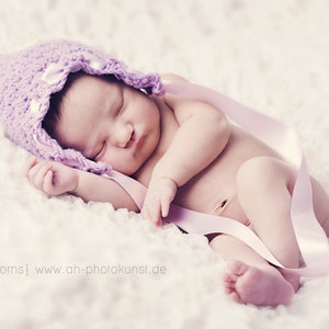 May include: A sleeping baby wearing a lavender crocheted bonnet with ribbon accents. The baby is lying on a soft, white, textured surface, with a pink ribbon draped across the body. Text on the image reads "{newborns} www.ah-photokunst.de".