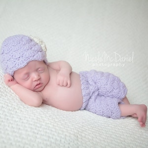 Puede incluir: Un bebé durmiendo con un gorro y pantalones de crochet color lavanda. El bebé está acostado sobre una manta de crochet blanca.
