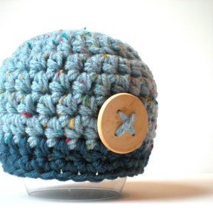 Puede incluir: Un gorro de punto azul y turquesa con un botón de madera con un diseño de cruz.