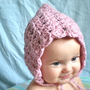 May include: A pink crocheted baby bonnet with a scalloped edge. The bonnet is worn by a baby with light skin and brown hair.