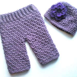 Puede incluir: Un conjunto de ropa de bebé de ganchillo en color morado. El conjunto incluye un pantalón y un gorro con una flor morada en la parte superior.