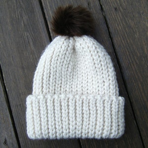 May include: A white knitted beanie with a brown faux fur pom pom on top.