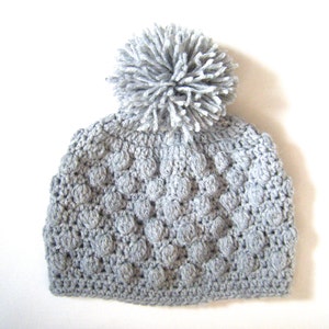 Könnte beinhalten: Eine graue Häkelmütze mit einem flauschigen Bommel oben. Die Mütze hat ein strukturiertes, unebenes Design.