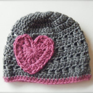May include: Gray crocheted beanie with a pink crocheted heart on the front. The beanie has a pink crocheted brim.