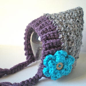 May include: Purple and grey crocheted baby hat with a blue flower and a button. The hat has a long tie.