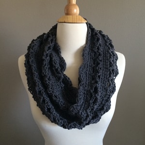 Könnte beinhalten: Ein grauer gehäkelter Infinity-Schal mit einem Spitzenmuster. Der Schal wird auf einer Holzpuppe präsentiert.