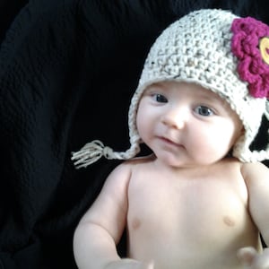 Könnte beinhalten: Ein Baby trägt eine gehäkelte Mütze mit einer rosa Blume oben. Die Mütze ist beige und hat einen weißen Bommel am Ende der Ohrenklappen.