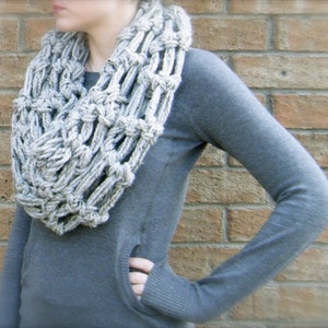 May include: A light gray, chunky knit infinity scarf. The scarf is made of a thick, textured yarn and has a large, open weave design. The person in the image is wearing a gray sweater with a pocket.