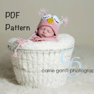 May include: A pink and white crocheted owl hat with a yellow beak and black eyes. The hat is worn by a baby sleeping in a white wicker basket. The text "PDF Pattern" is written in black at the top of the image.