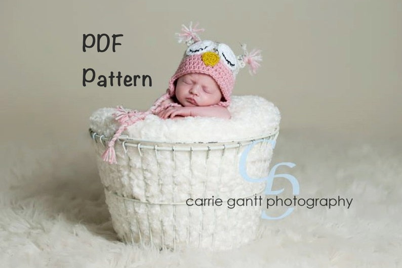 May include: A pink and white crocheted owl hat with a yellow beak and black eyes. The hat is worn by a baby sleeping in a white wicker basket. The text "PDF Pattern" is written in black at the top of the image.
