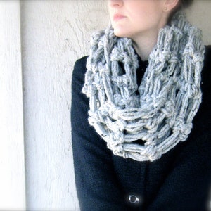 May include: A gray crocheted infinity scarf with a loose, open weave pattern. The scarf is worn by a person with a black jacket.