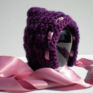 May include: Purple crocheted hat with pink satin ribbon ties. The hat is on a mannequin head.