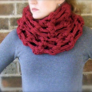 May include: A red knitted infinity scarf with a chunky, open-weave design. The scarf is worn by a person with a gray sweater.