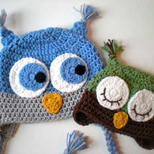 May include: Two handmade crochet owl hats. One is blue, gray, and white with a yellow beak. The other is brown, green, and white with a yellow beak. Both have large, round eyes and ear tufts.