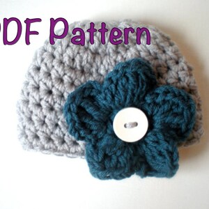 Puede incluir: Un gorro de ganchillo gris con una flor verde azulado y un botón blanco en el centro. El texto "PDF Pattern" está en morado encima del gorro. El gorro está hecho a mano y es adecuado para bebés o niños.