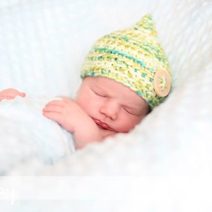 Peut inclure: Un nouveau-né dort paisiblement dans une couverture blanche, portant un bonnet en tricot jaune et vert avec un bouton.