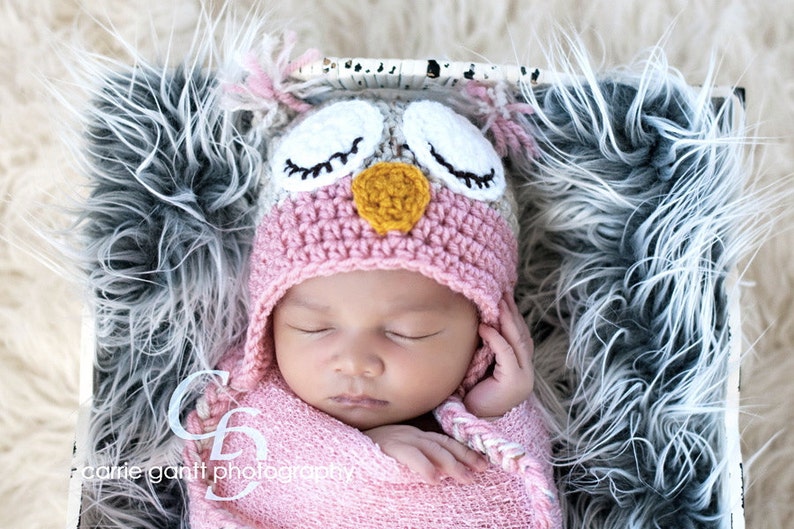 May include: A newborn baby wearing a pink and brown crocheted owl hat with white fur trim. The baby is sleeping peacefully in a white and gray fur-lined basket.