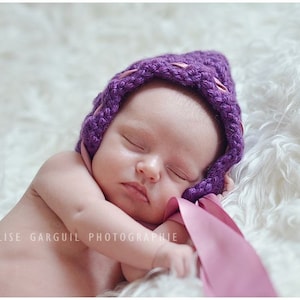 May include: A sleeping baby wearing a purple crocheted bonnet with a pink ribbon tied around the chin.