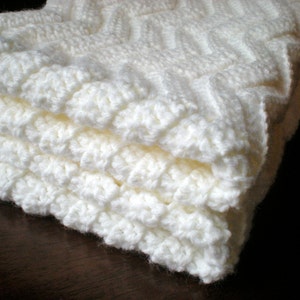 May include: A stack of folded, off-white, crocheted blankets. The blankets have a textured, raised pattern, and are made of a soft, fluffy yarn. The blankets are neatly stacked, showing the detailed stitch work.