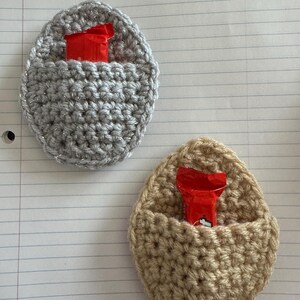 May include: Two crocheted egg-shaped pouches, one silver and one beige, each containing a small red candy bar.