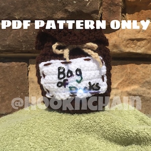 May include: A crocheted brown bag with a white label that says "Bag of Tricks" and a tan string tied around the top. The bag is sitting on a green towel.