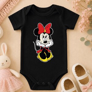 A black baby onesie with a Minnie Mouse design, a pair of white baby shoes, and a stuffed bunny rabbit are arranged on a beige surface.