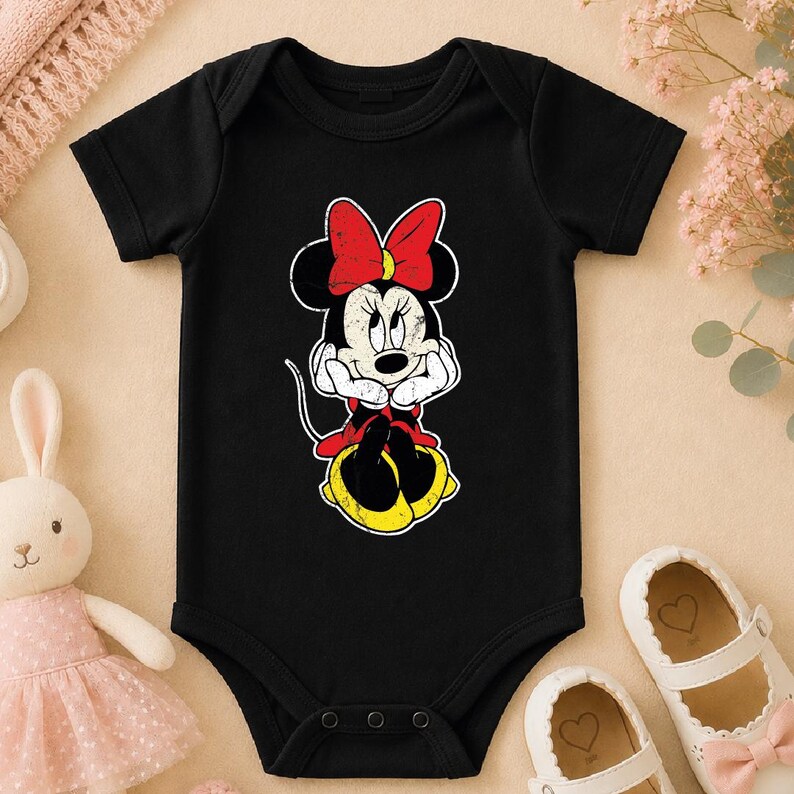 A black baby onesie with a Minnie Mouse design, a pair of white baby shoes, and a stuffed bunny rabbit are arranged on a beige surface.