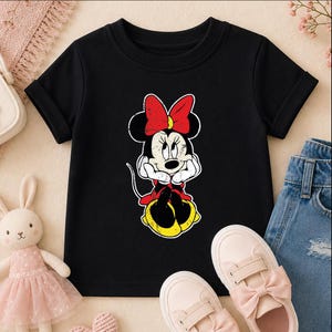 A black t-shirt with a Minnie Mouse graphic, a pair of pink sneakers, and a pair of blue jeans are arranged on a surface.