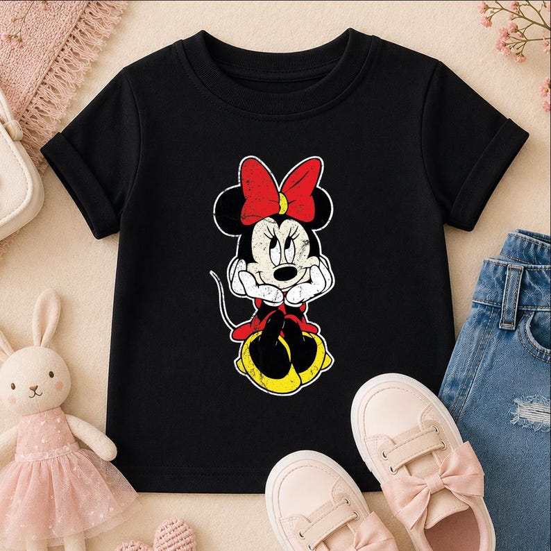 A black t-shirt with a Minnie Mouse graphic, a pair of pink sneakers, and a pair of blue jeans are arranged on a surface.