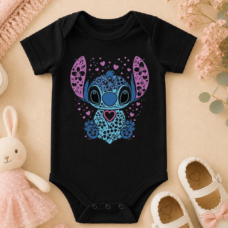 A black baby onesie with a colorful design featuring a cartoon character, surrounded by pink and white baby shoes and a stuffed bunny.