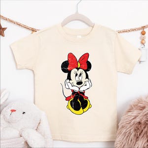 A white t-shirt with a Minnie Mouse graphic printed on it, hanging on a wooden hanger.