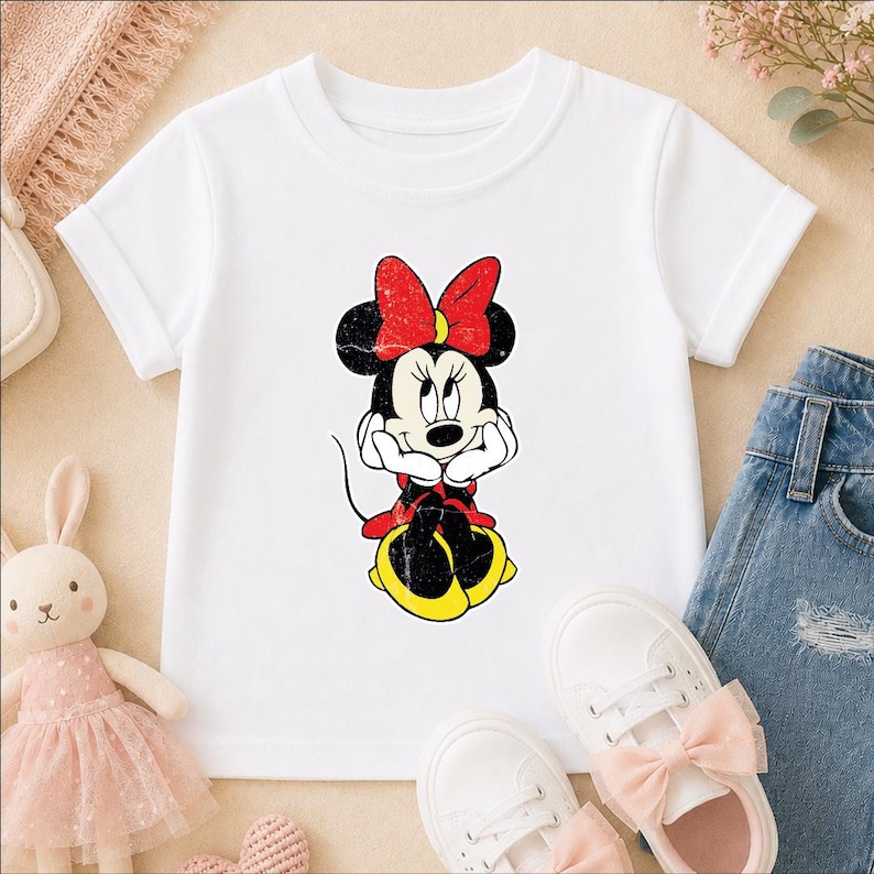 A white t-shirt with a Minnie Mouse graphic, a pair of white sneakers, and a pair of blue jeans are arranged on a surface.