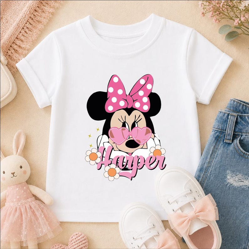A white t-shirt with a Minnie Mouse design and the name "Hacer" printed on it, along with pink polka dots and flowers. The t-shirt is placed on a beige surface, and there are pink shoes and a stuffed bunny rabbit nearby.