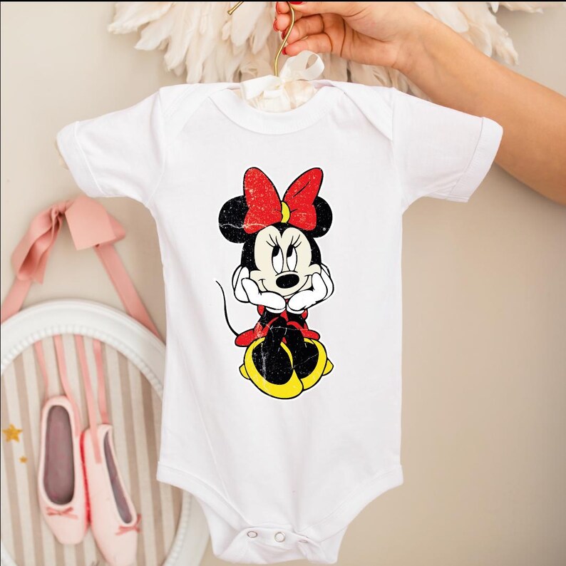 A white baby onesie with a Minnie Mouse design is being held by a person's hand.