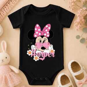 A black baby onesie with a Minnie Mouse design and the name "Hester" printed on it, along with pink polka dots and flowers. The onesie is placed on a beige surface, and there are a pair of white shoes and a stuffed bunny rabbit nearby.