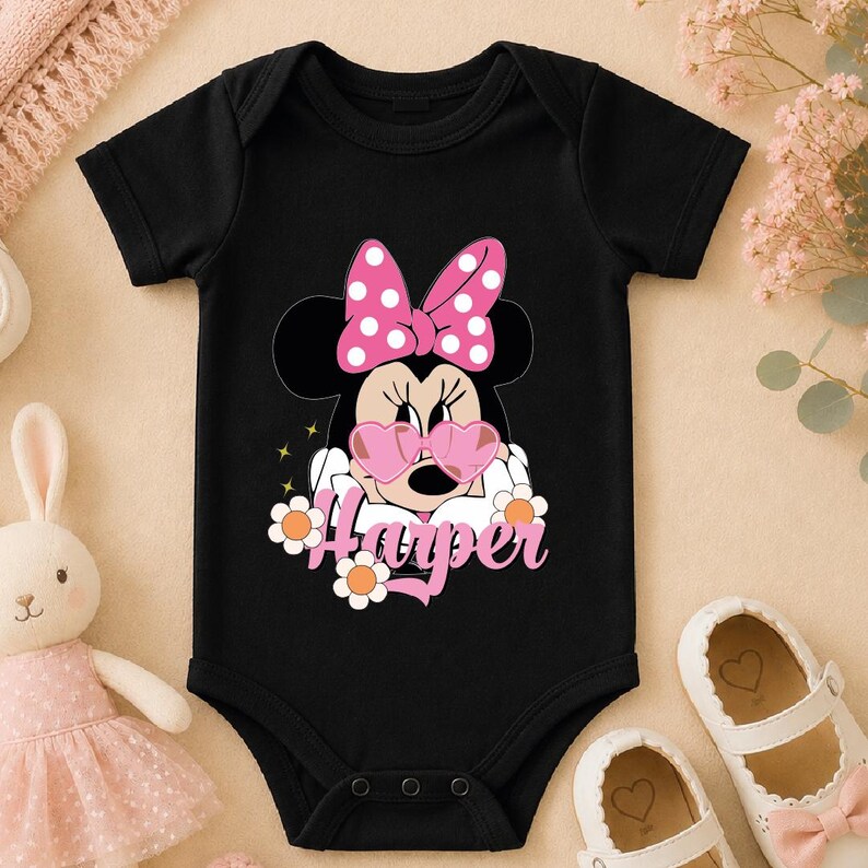 A black baby onesie with a Minnie Mouse design and the name "Hester" printed on it, along with pink polka dots and flowers. The onesie is placed on a beige surface, and there are a pair of white shoes and a stuffed bunny rabbit nearby.