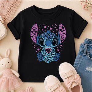 A black t-shirt with a colorful graphic of a cartoon character, surrounded by pink and blue flowers, hearts, and bows. The t-shirt is placed on a beige surface, with a pair of pink and blue sneakers and a stuffed bunny rabbit nearby.