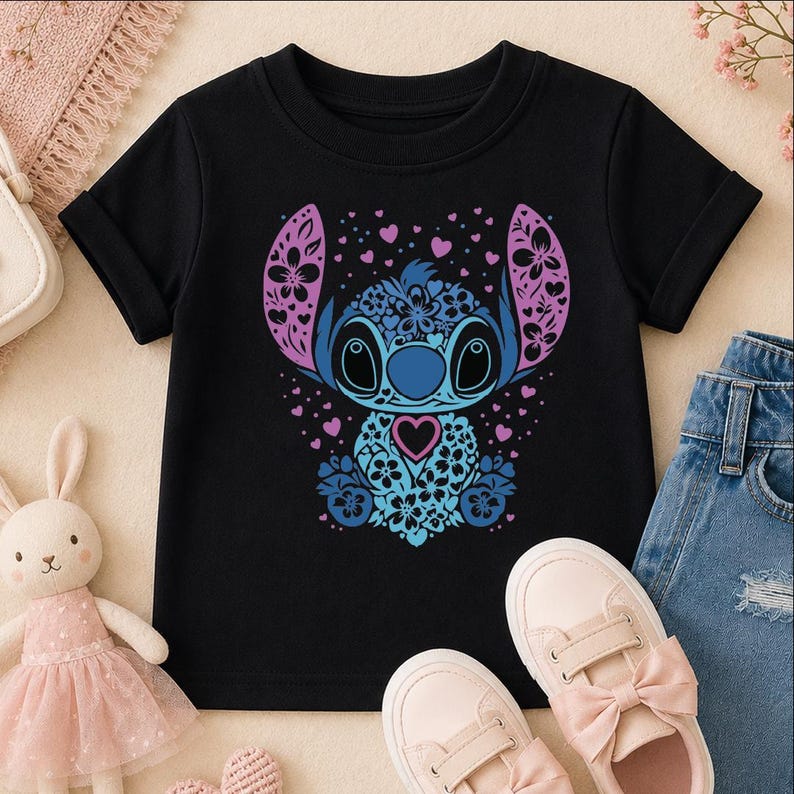 A black t-shirt with a colorful graphic of a cartoon character, surrounded by pink and blue flowers, hearts, and bows. The t-shirt is placed on a beige surface, with a pair of pink and blue sneakers and a stuffed bunny rabbit nearby.