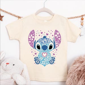 A white t-shirt with a colorful graphic design featuring a cartoon character with large eyes, floral patterns, and a heart shape.