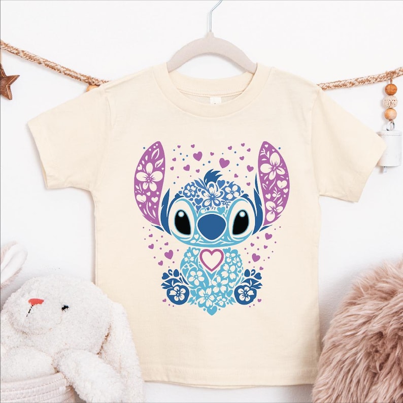 A white t-shirt with a colorful graphic design featuring a cartoon character with large eyes, floral patterns, and a heart shape.