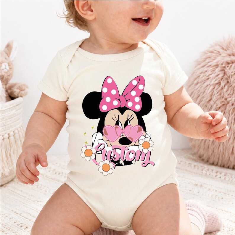 A baby wearing a white onesie with a Minnie Mouse design and the word "Pritom" printed on it.