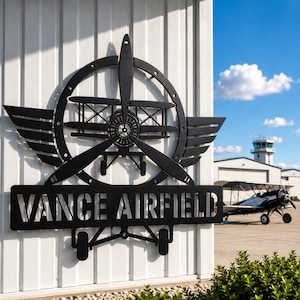 May include: A black metal wall sign featuring a biplane silhouette centered within a circular frame with wings, mounted on a white vertical panel. The sign displays the text VANCE AIRFIELD in bold, cut-out capital letters at the bottom.